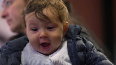 Annenin kollarında bebek, ağzı açık, heyecanlı ifade, neşeli an, günlük aile sahnesi, kışlık ceket, mutlu bebeğin yakın çekimi, neşeli etkileşim