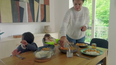 Anne yemek masasında çoklu görevdeyken, bebek yüksek sandalyede otururken ve çocuk tabağına odaklanmışken, aile yemeğine hazır.
