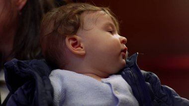 Bebek annesinin kollarında, yukarı bakıyor, rahat bir ceket giyiyor, düşünceli bir ifade takınıyor, günlük aile anları, çevreyi gözlemliyor, yakın çekim manzarası, ev ortamları...