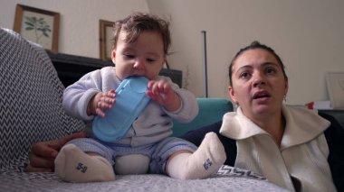 Bebek koltukta oturup mavi nesneyi çiğniyor, anne gözlemliyor, evde huzurlu bir aile anı yaşıyor, bebek oynuyor ve keşfediyor, bebekle anne arasında bağ kuruyor, oturma odası kuruyor.