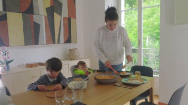 Anne yemek masasında yemek servisi yaparken, büyük çocuk ve bebek bakıcılığı, aile yemeği hazırlama ve sıcak bir evde toplanma.