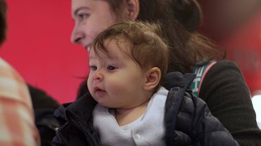 Annenin kollarındaki bebek, özenle bakıyor, günlük aile anı, ceket giyiyor, açık havada ya da kapalı alanda, samimi etkileşim, ebeveyn-çocuk bağı, çocuk gözetleme ortamı.