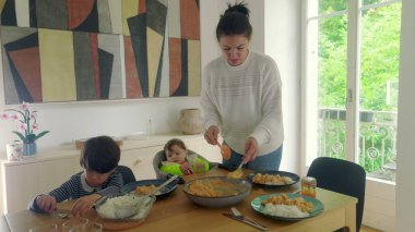 Anne yemek masasında çoklu görevdeyken, bebek yüksek sandalyede otururken ve çocuk tabağına odaklanmışken, aile yemeğine hazır.
