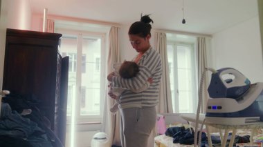 Anne bebeğini emzirirken huzurlu bir ev ortamında duruyor, sessiz beslenme anı, yakın aile bağı, anne bakımı ve evde şefkat.