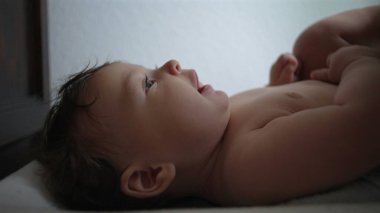 Bebek sırt üstü uzanıyor, yan profil, nişanlı yüz ifadesi, kollar karnında dinleniyor, sakin an, kapalı alan, ev ortamı, yakın görüş, yumuşak bebek teni, huzurlu bakışlar.