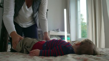 Anne bebek elbiselerini değiştirirken bebek yatakta yatar, hassas ve dikkatli bir şekilde bakım yapar, ev ortamında bebek hijyenine ve hijyene dikkat eder..