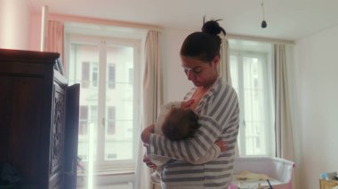 Anne, bebeğini yatak odasında durup emzirirken huzur dolu bir yuva ortamında huzur verici, şefkatli bir an yaşıyor.