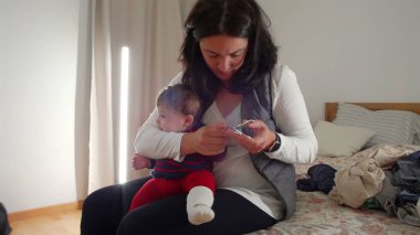 Anne yatakta oturup bebek tırnağı kesiyor, gerekli bakım ve bakımı sağlıyor, sıcak bir ev ortamında samimi ebeveynlik anları yaşıyor, bebek hijyenine odaklanıyor..