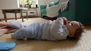 Bebek ağzında parmaklarıyla sırt üstü uzanıyor, sakin ve rahat bir yüz ifadesi, evde aile zamanı, bebek gezisi çevresi, huzurlu bir an, rahat bir evde neşeli bir çocuk.