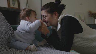 Anne bebekle kanepede iletişim kuruyor, şakacı yüz yüze iletişim kuruyor, erken çocukluk gelişiminde neşe ve bağlılık gösteriyor, ev ortamını büyütüyor.