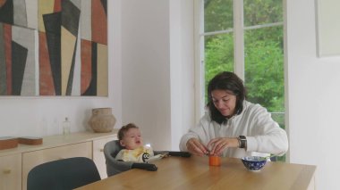 Anne yemek masasında bebeği emziriyor, bebek beslenme sırasında rahatsızlığını dile getiriyor, anne nazikçe yemek yemesi için teşvik etmeye çalışıyor, yemek sırasında etkileşimi besliyor.