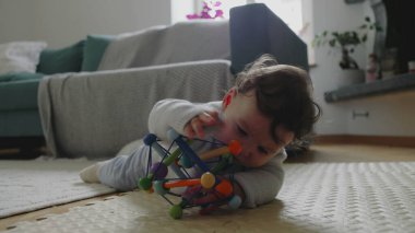 Bebek paspasın üzerinde yatıyor, iki eliyle renkli oyuncakları tutuyor ve güvenli bir ev ortamında erken gelişim sırasında şekilleri ve dokuları keşfediyor.