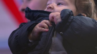 Bebek ceketi çiğniyor, yakından bakıyor, tuhaf bir ifade takınıyor, kışlık ceket giyiyor, günlük aile anı, beslenme ve koruyucu sahne, ev ortamında rahat.