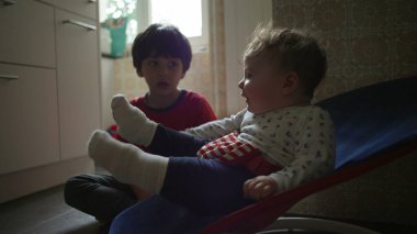 Bebek bir fedainin içinde ağabeyi izlerken yukarı bakıyor, sakin bir sabah rutini sırasında sıcak bir aile mutfağında tuhaf bir kardeş etkileşimi.