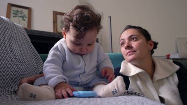 Bebek koltukta oturup mavi nesneyi çiğniyor, anne gözlemliyor, evde huzurlu bir aile anı yaşıyor, bebek oynuyor ve keşfediyor, bebekle anne arasında bağ kuruyor, oturma odası kuruyor.