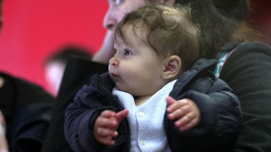 Annenin kollarında bir bebek, ceketli, heyecanlı bir yüz ifadesi, samimi bir ilişki, günlük aile anı, çevreyi gözlemleme, yakın görüş.