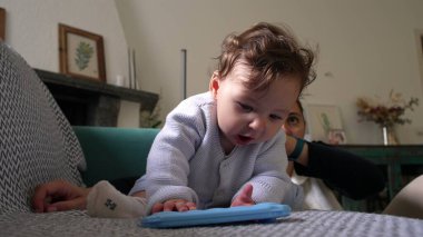 Bebek, mavi nesneyle oynarken koltukta öne eğiliyor, odaklanmış ifade, merakla bebek keşif nesnesi, kapalı alanda eğlenceli bir an, yakınlarda annesiyle aile bağı kurma zamanı.