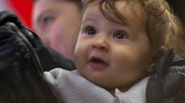 Bebek annesinin kollarında, açık gözlerle bakıyor, ceket giyiyor, tuhaf bir ifade takınıyor, günlük aile anı, yakın çekim görüntüsü, kapalı mekanda samimi etkileşim.