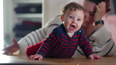 Bebek gülümsüyor ve masada emekliyor, neşeli ve enerjik, anne arka planda izliyor, bebek ve anne arasında canlı bir etkileşim, bebek anın tadını çıkarıyor.