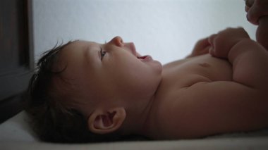 Bebek sırt üstü uzanıyor, yan profil, nişanlı yüz ifadesi, kollar karnında dinleniyor, sakin an, kapalı alan, ev ortamı, yakın görüş, yumuşak bebek teni, huzurlu bakışlar
