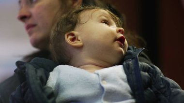 Bebek annesinin kollarında, yukarı bakıyor, rahat bir ceket giyiyor, tuhaf bir ifade takınıyor, günlük aile anları, çevreyi gözlemliyor, yakın çekim manzarası, günlük ev ortamları.