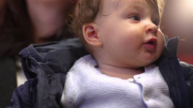 Bebek annesinin kollarında, usulca gülümsüyor, ceketini giyiyor, neşeli bir ifade takınıyor, samimi bir aile anı, çevreyi gözlemliyor, yakın çekim, gündelik etkileşim, sıcaklık ve şefkat.