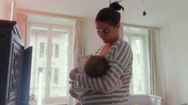 Anne, bebeğini yatak odasında durup emzirirken huzur dolu bir yuva ortamında huzur verici, şefkatli bir an yaşıyor.