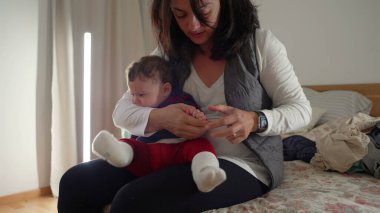 Anne bebek tırnaklarını keserken yatakta oturuyor, bebek hijyenine bakıyor, evde birden fazla iş yapıyor, gerekli özen ve ilgiyi sağlıyor..