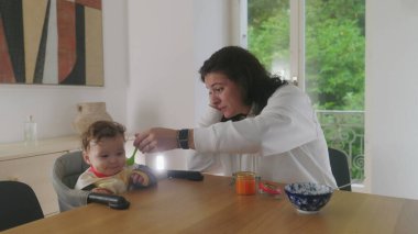 Anne yemek masasında bebeği besliyor, bebek heyecanla gülümsüyor, anne ve bebek arasındaki neşeli etkileşim, neşeli beslenme anı, aile yemeği, bebeğin yemekten zevk alması.