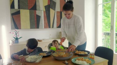 Anne yemek masasında çoklu görevdeyken, bebek yüksek sandalyede otururken ve çocuk tabağına odaklanmışken, aile yemeğine hazır.