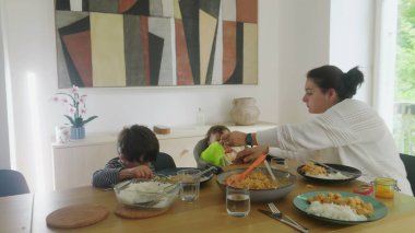 Anne bebeğini beslerken büyük çocuk kendi yemeğine odaklanıyor, aile yemek masasında birlikte yemek yiyor, evde ebeveynlik yapıyor, bebek yüksek sandalyede yiyor, bağ kuruyor.