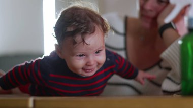 Bebek masada gülüyor ve emekliyor, heyecan ve enerji, anne arka planda gözlem yapıyor, neşeli etkileşim, neşeli bebek ev içi aile zamanlarında coşku dolu.