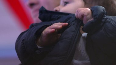 Bebek ceketi çiğniyor, yakından bakıyor, tuhaf bir ifade takınıyor, kışlık ceket giyiyor, günlük aile anı, beslenme ve koruyucu sahne, ev ortamında rahat.