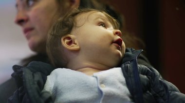 Bebek annesinin kollarında, yukarı bakıyor, rahat bir ceket giyiyor, tuhaf bir ifade takınıyor, günlük aile anları, çevreyi gözlemliyor, yakın çekim manzarası, günlük ev ortamları