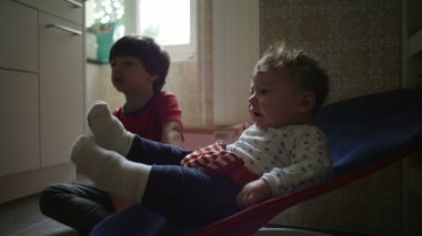 Bebek bir fedainin içinde ağabeyi izlerken yukarı bakıyor, sakin bir sabah rutini sırasında sıcak bir aile mutfağında tuhaf bir kardeş etkileşimi.