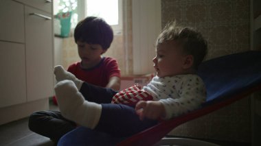 Bebek bir fedainin içinde ağabeyi izlerken yukarı bakıyor, sakin bir sabah rutini sırasında sıcak bir aile mutfağında tuhaf bir kardeş etkileşimi.