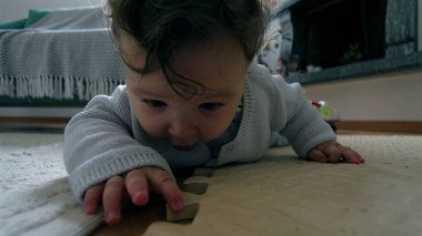 Bebek yerde yatıyor, yüzü yere yakın, tahta bir blokla etkileşimde, merakla çevreyi keşfediyor, ev ortamında güzel motor beceriler geliştiriyor.