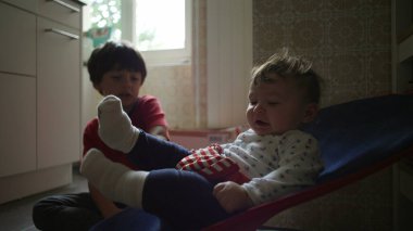 Bebek bir fedainin içinde ağabeyi izlerken yukarı bakıyor, sakin bir sabah rutini sırasında sıcak bir aile mutfağında tuhaf bir kardeş etkileşimi.