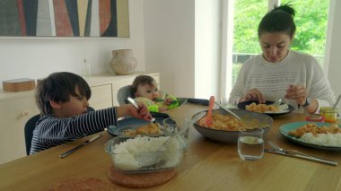Anne yemek masasında servis yapıyor, bebek yüksek sandalyede ve çocuk yemek yiyor, aile yemeği sıcak bir evde.