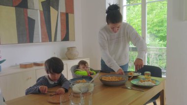 Anne yemek masasında yemek servisi yaparken, büyük çocuk ve bebek bakıcılığı, aile yemeği hazırlama ve sıcak bir evde toplanma.