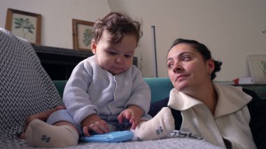 Bebek koltukta oturup mavi nesneyi çiğniyor, anne gözlemliyor, evde huzurlu bir aile anı yaşıyor, bebek oynuyor ve keşfediyor, bebekle anne arasında bağ kuruyor, oturma odası kuruyor.