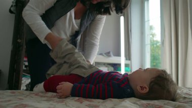 Anne bebek elbiselerini değiştirirken bebek yatakta yatar, hassas ve dikkatli bir şekilde bakım yapar, ev ortamında bebek hijyenine ve hijyene dikkat eder..