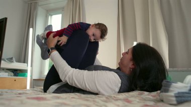 Anne yatakta yatarken bebeği kaldırıyor, neşeli ve eğlenceli etkileşimler kuruyor, aile evinde birlikte vakit geçiriyor ve eğleniyor, çocukla mutlu ve samimi bir an.