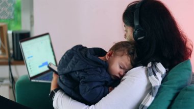 Anne kucağında uyuyan bir bebeği tutarken telefonunu kontrol eder ve dizüstü bilgisayarda çalışır, bakım ve uzaktan işleri harmanlar, ev ortamında birden fazla iş yapar.