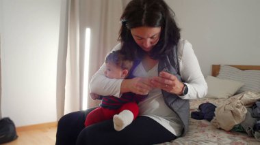 Anne yatakta oturup bebek tırnağı kesiyor, gerekli bakım ve bakımı sağlıyor, sıcak bir ev ortamında samimi ebeveynlik anları yaşıyor, bebek hijyenine odaklanıyor..