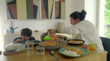Anne yemek masasında servis yapıyor, bebek yüksek sandalyede ve çocuk yemek yiyor, aile yemeği sıcak bir evde.