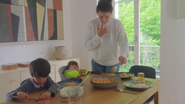 Anne yemek masasında yemek servisi yaparken, büyük çocuk ve bebek bakıcılığı, aile yemeği hazırlama ve sıcak bir evde toplanma.