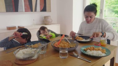 Anne yemek masasında bebeği beslerken büyük çocuk yemek yiyor, aile yemeği yiyor, ev içinde huzurlu bir ortam sağlıyor, anne çoklu görev yapıyor, yüksek sandalyedeki bebek, aile bakımı...