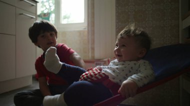 Bebek bir fedainin içinde ağabeyi izlerken yukarı bakıyor, sakin bir sabah rutini sırasında sıcak bir aile mutfağında tuhaf bir kardeş etkileşimi.
