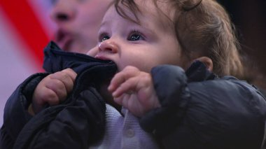 Bebek ceketi çiğniyor, yakından bakıyor, tuhaf bir ifade takınıyor, kışlık ceket giyiyor, günlük aile anı, beslenme ve koruyucu sahne, ev ortamında rahat.